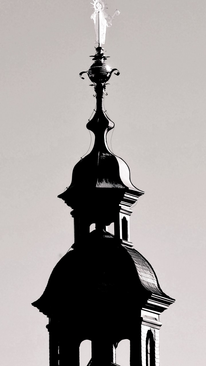 Das Foto in schwarz/weiss zeigt eine dunkle Turmspitze der Stiftskirche mit heller, spiegelnder Figur an der Spitze.