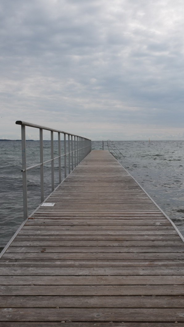 Das Foto zeigt einen Holzsteg mit Stahlgeländer das ins Meer führt. Bei stark bewölktem Himmel und leicht welligem Meer. Aufgenommen im August 2023 in der Region Mitteljütland, Dänemark